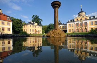 Historisches Gebäudeensemble mit gelber Fassade, klassischer Architektur, Brunnen und Spiegelung im Wasser bei klarem Himmel